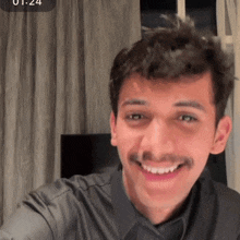 a man with a mustache is smiling in front of a window with the time 1:24