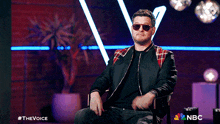 a man wearing sunglasses and a plaid jacket is sitting in a chair with a nbc logo in the background