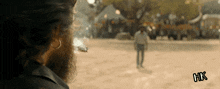 a man with a beard is smoking a cigarette in a field with hk written on the bottom right