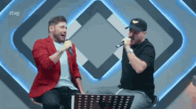 two men singing into microphones in front of a rtve sign