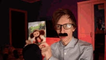 a man with glasses and a fake mustache holds a picture of a stuffed monkey