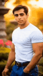 a man wearing a white t-shirt and blue jeans is standing in front of a fountain with his hands in his pockets .