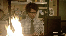 a man in a plaid shirt and tie is typing on a computer with flames coming out of the monitor