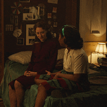 two women sit on a bed in a dark room with pictures on the wall