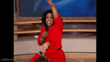 oprah winfrey is sitting on a stage holding a microphone and raising her arms in the air .