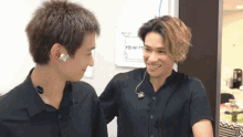 two men are standing next to each other and one has a sign on the wall that says kiss-m-ft