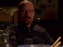 a bald man with glasses and a beard is sitting at a table with a bowl of food .