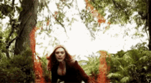 a woman with red hair is standing in a forest surrounded by trees and a red circle of fire .