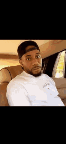 a man with a beard is sitting in the back seat of a car wearing a hat and a white shirt .