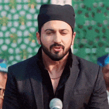 a man with a beard wearing a black hat and a black suit is standing in front of a microphone .