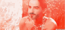 a shirtless man with a beard and a necklace around his neck is sitting in front of a red background .