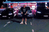a man in a mercedes shirt is standing in front of three cars in a parking lot .