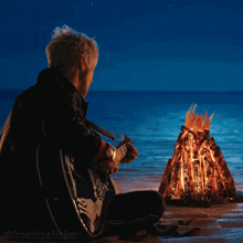 a man sits on the beach playing a guitar next to a small fire
