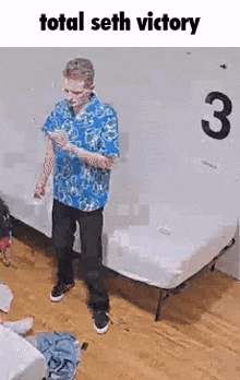 a man in a blue shirt is standing next to a bed in a room with a sign that says total seth victory .