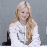 a woman with blonde hair is sitting at a table wearing a white hoodie and smiling .