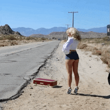 a woman standing on the side of a road next to a suitcase that says ' triumph ' on the handle