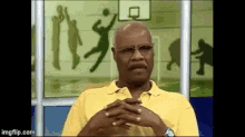 a man wearing glasses and a yellow shirt is sitting with his hands folded in front of a basketball hoop .