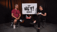 two men sitting in front of a sign that says hey