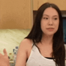 a woman is standing in a kitchen wearing a white tank top and looking at the camera .