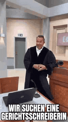 a man in a judge 's robe is standing in a courtroom in front of a laptop computer .