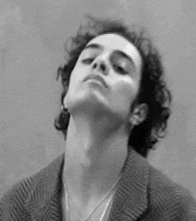 a black and white photo of a man with curly hair wearing a jacket and necklace .