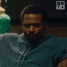 a man in a blue sweater is sitting on a couch with a be t logo behind him