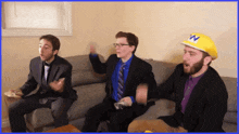 three men are sitting on a couch with one wearing a yellow w hat
