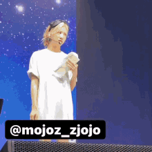a woman in a white dress stands in front of a microphone and a sign that says @mojoz_zjojo on it