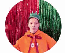 a man wearing an orange hoodie and a birthday crown is standing in front of a red and green curtain .