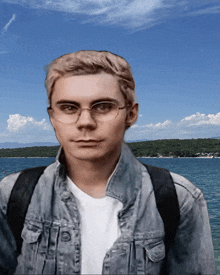 a man wearing glasses and a denim jacket stands in front of a lake