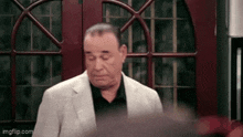 a man in a suit and black shirt is standing in front of a red door and looking down .