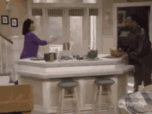 a woman in a purple shirt is standing in a kitchen talking to a man in a black jacket