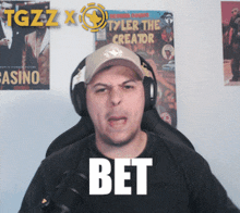 a man wearing headphones and a hat says bet in front of a poster for tyler the creator