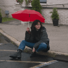 a woman holding a red umbrella sits on a curb
