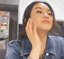 a woman in a denim jacket is touching her face in front of a fridge .
