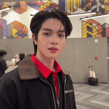 a young man wearing a black jacket and a red shirt is standing in front of a wall .