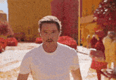 a man with a mustache wearing a white t-shirt stands in front of a crowd of people