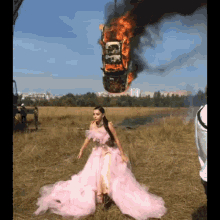 a woman in a pink dress is standing in a field with a burning car in the background