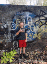 a young boy giving a thumbs up in front of a wall covered in graffiti