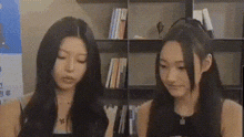 two women are sitting next to each other in front of a bookshelf in a library .
