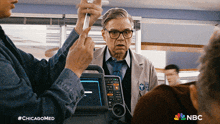 a man in a lab coat is talking to a group of people with the nbc logo in the background