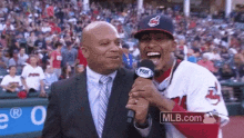 a man in a suit and tie is talking into a microphone while a baseball player in an indians uniform laughs