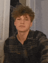 a young man in a plaid shirt looks at the camera with a serious look on his face