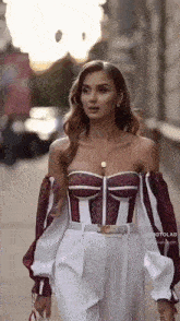 a woman is walking down a street wearing a burgundy and white off the shoulder top and white pants .