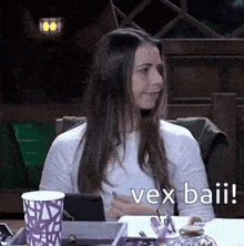 a woman is sitting at a table with a cup of coffee and a cell phone and says vex baii .