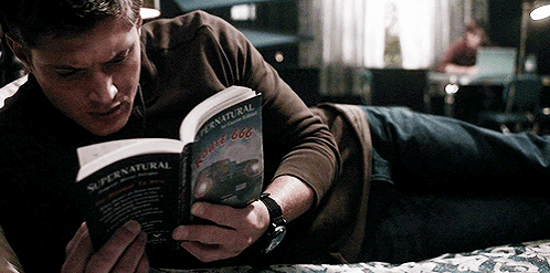 A man lies on a bed reading the book Supernatural. It's Dean Winchester, from Supernatural