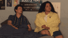 a man and woman sit on a bed in front of a poster for sous le vent