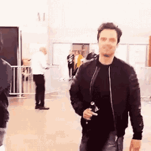 a man in a black jacket is walking in a hallway holding a bottle of water .