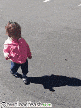 a little girl in a pink jacket is walking down the street and her shadow is on the ground .