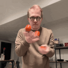 a man wearing glasses and a brown sweater holds two orange balls in his hands